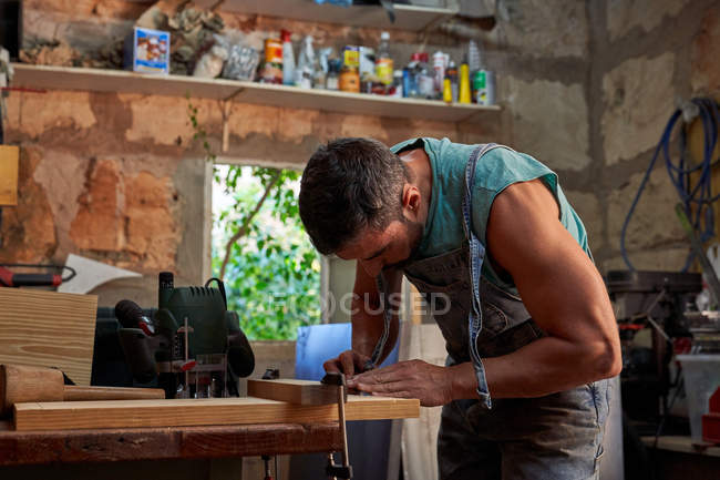 Side view of skilled middle aged handyman in overalls using clamp while working with wooden plank in workshop — Stock Photo