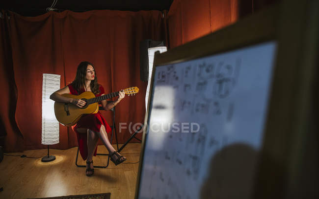 Affascinante donna artistica in abito rosso che suona la canzone alla chitarra sul palco con luce calda in Spagna — Foto stock