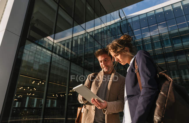 Оптимистичные бизнесмены улыбаются и просматривают планшеты, стоя снаружи современного здания и отдыхая после работы — стоковое фото