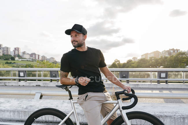 Happy adult bearded man in black cap wearing black shirt and beige shorts resting with bicycle across footbridge in city looking away — Stock Photo