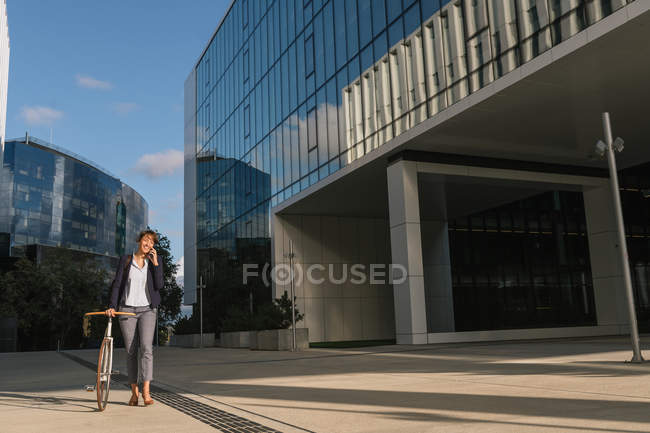 Positive Geschäftsfrau mit Fahrrad lächelt und spricht auf dem Smartphone, während sie an einem sonnigen Tag auf der Stadtstraße vor einem modernen Gebäude spaziert — Stockfoto