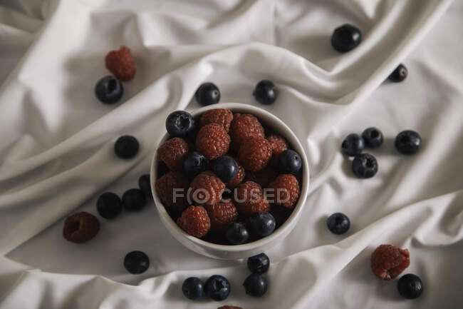 Draufsicht auf schmackhafte Himbeeren und Blaubeeren im Teller auf weißem Seidenstoff — Stockfoto