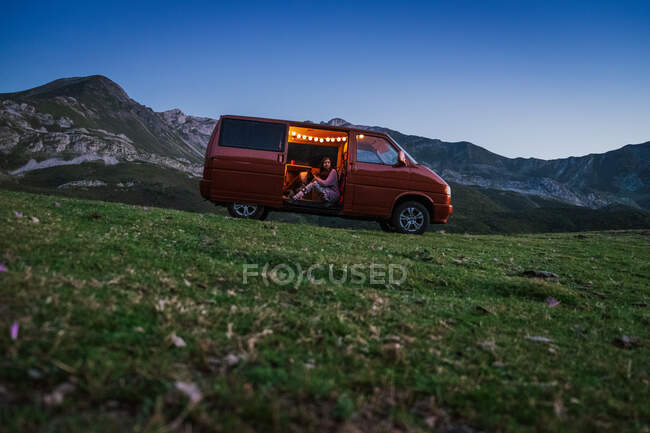 Woman with Border Collie dog sitting inside of red van against rocky mountains under cloudless blue sky in summer evening — Stock Photo