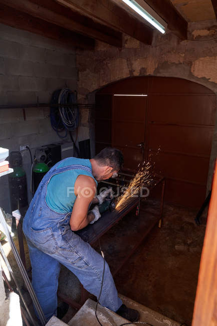Seitenansicht des Arbeiters mit Schutzbrille und Handschuhen beim Schneiden von Metall mit einem Schleifer mit Funkenflug während der Arbeit in der Werkstatt — Stockfoto