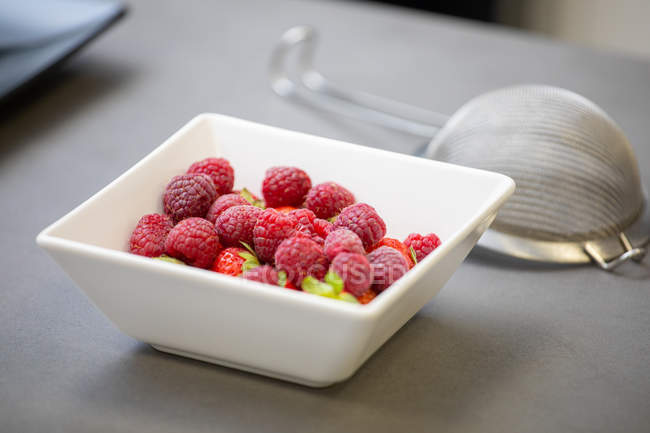 Fragrant appetizing fresh raspberries with green stem in white square bowl and metal sieve with handle on table — Stock Photo