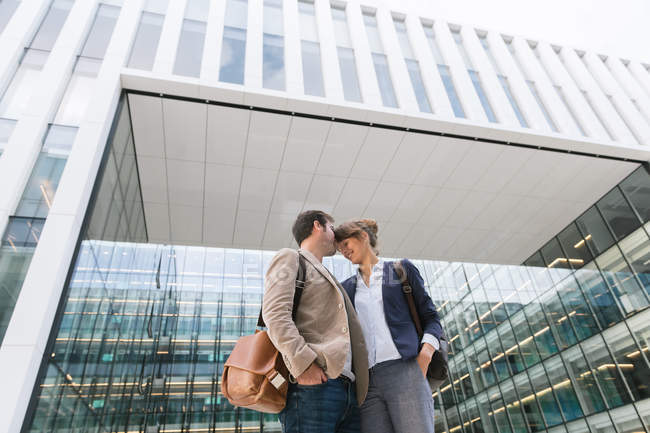 Von unten glückliche Paar-Kollegen, die sich küssen, während sie nach Feierabend vor einem modernen Gebäude an der Stadtstraße stehen — Stockfoto