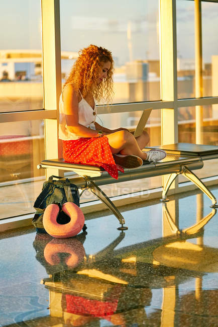 Femme élégante gaie tapant sur ordinateur portable tout en étant assis avec les jambes croisées sur un banc en métal dans le couloir en verre de l'aéroport au Texas — Photo de stock