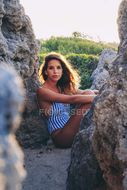 Woman in stylish striped swimsuit having rest among rocks and looking at camera with green fields on background — Stock Photo