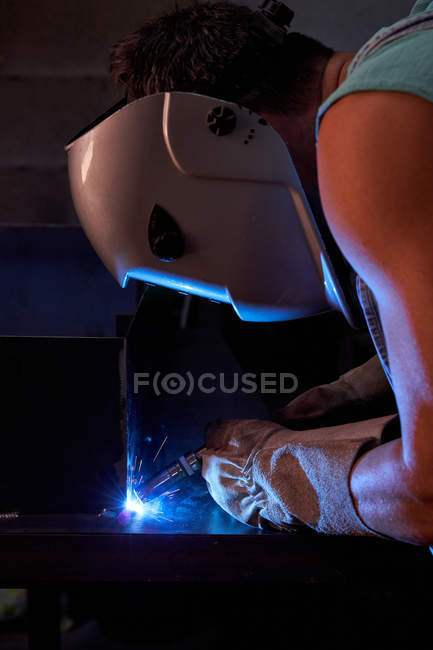Seitenansicht des Handwerkers in Schutzmaske mit Overalls und Handschuhen bei der Arbeit in der Werkstatt und beim Löten von Metall — Stockfoto