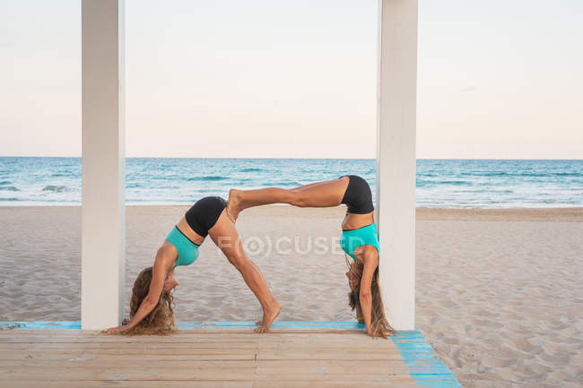 Side view of slim active women performing corner on dog down together in acroyoga on wooden stand on seaside — Stock Photo