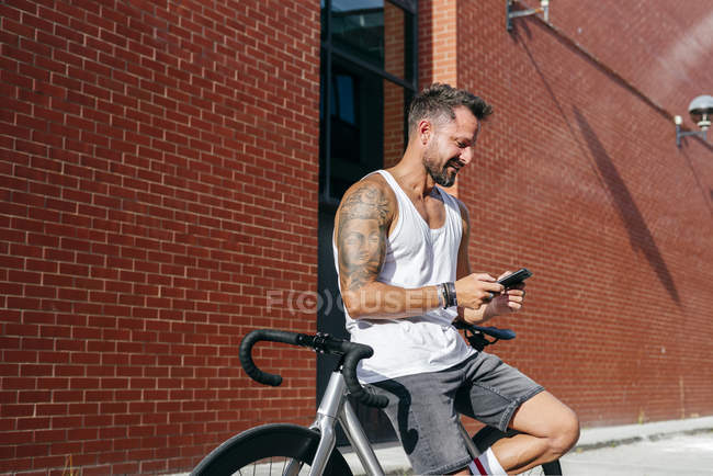Schöner männlicher Radfahrer in Sportkleidung mit Smartphone, während er auf dem Fahrrad neben roter Backsteinmauer ruht — Stockfoto