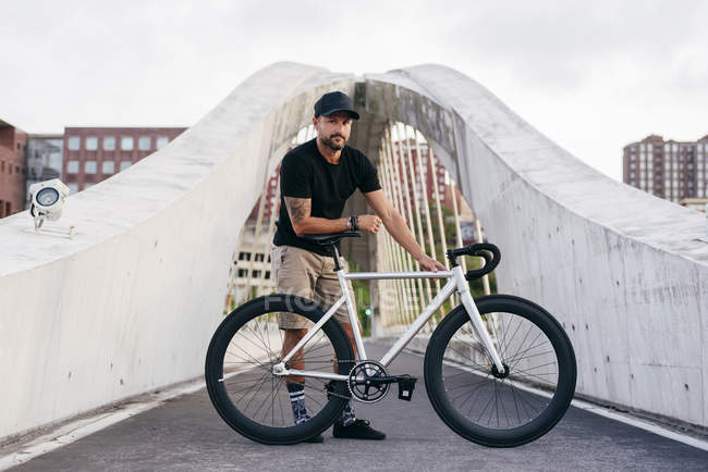 Happy adult bearded man in black cap wearing black shirt and beige shorts standing with bike across footbridge in city looking at camera — Stock Photo
