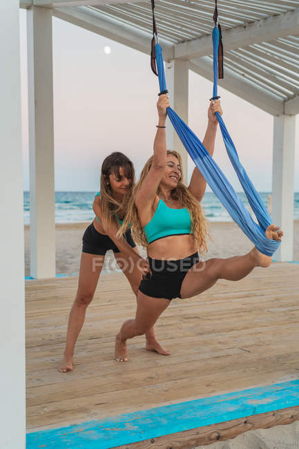 Careful woman supporting girl friend making acroyoga exercise hanging on hammock on wooden stand at beach — Stock Photo