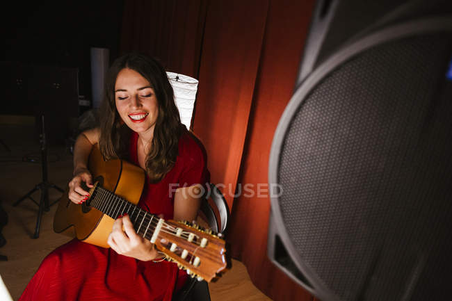 Обаятельная артистка в красном платье исполняет песню, играя на гитаре на сцене с теплым светом в Испании — стоковое фото