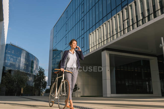 Позитивна бізнес-леді з велосипедом посміхається і виступає на смартфоні під час виходу за межі сучасної будівлі в сонячний день на міській вулиці — стокове фото