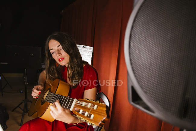 Приваблива жінка з заплющеними очима в червоному вбранні виконує пісню на гітарі на сцені з теплим світлом в Іспанії. — стокове фото