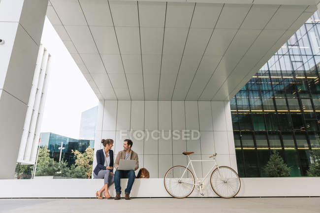 Delighted businesspeople smiling and browsing laptop together while sitting outside modern building near bicycle on city street — Stock Photo