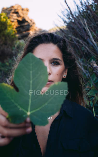 Mujer relajante en la naturaleza mientras cubre media cara con hoja verde de higo y mirando a la cámara - foto de stock