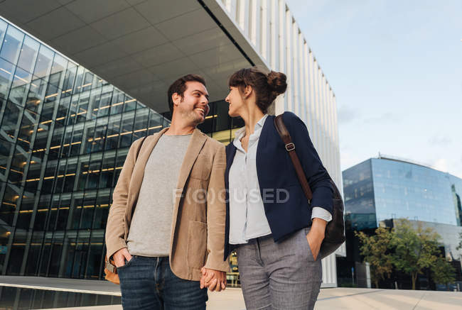 Desde abajo feliz pareja compañeros de trabajo en trajes casuales sonriendo y caminando juntos fuera de edificio moderno en la calle de la ciudad después del trabajo - foto de stock