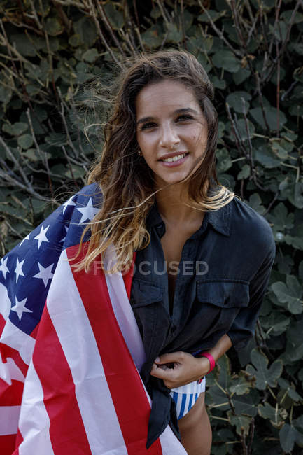 Femme en chemise noire décontractée et drapeau américain sur le dos regardant la caméra avec des plantes vertes sur fond — Photo de stock