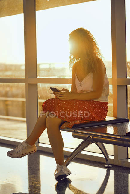 Agradable mujer alegre en auriculares escuchando música con teléfono móvil mientras se enfría en un banco de metal en el aeropuerto de Texas - foto de stock