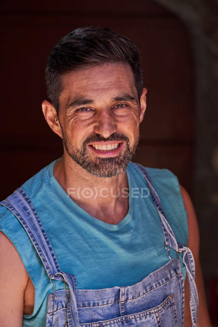 Joyeux travailleur barbu et souriant d'âge moyen portant une chemise bleue et des salopettes en jean avec un fond sombre — Photo de stock