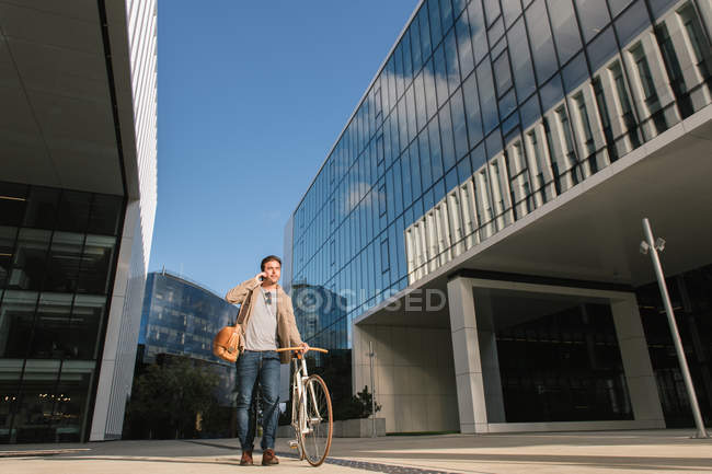 Людина в повсякденному одязі робить телефонний дзвінок, стоячи з велосипедом проти сучасного бізнес-центру висотного підйому зі скляними стінами в центрі міста — стокове фото