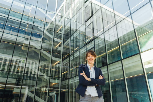 Positive female manager with crossed arms smiling while standing outside contemporary building with glass walls on city street — Stock Photo