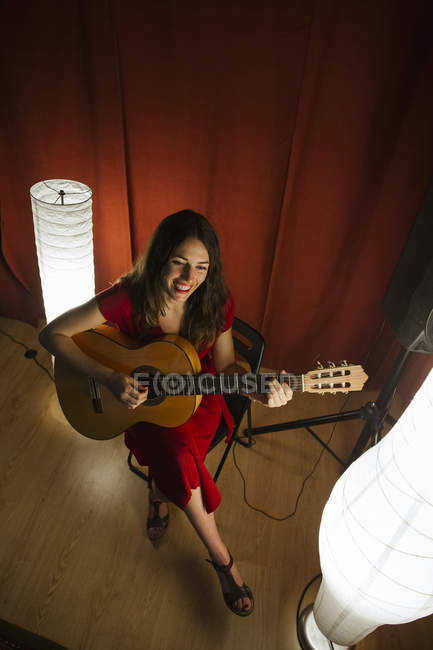 D'en haut femme talentueuse en robe rouge chantant et jouant de la guitare sur scène éclairée chaude lampe blanche à proximité — Photo de stock