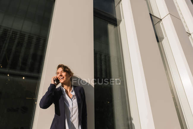 Mujer de negocios optimista con chaqueta oscura sonriendo y mirando hacia otro lado mientras se apoya en la pared del edificio en la calle de la ciudad y responde a la llamada telefónica - foto de stock