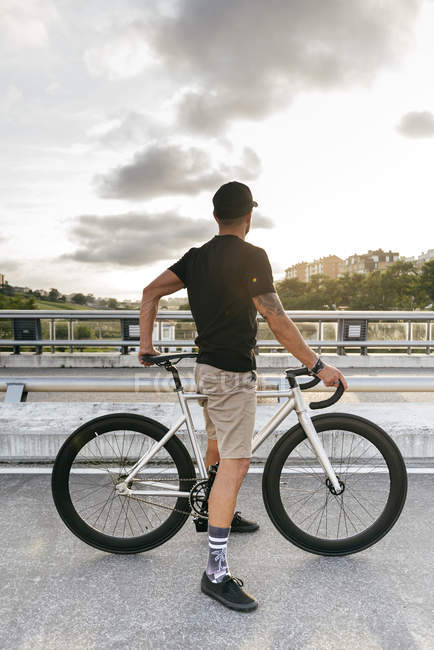 Vista posteriore dell'uomo in berretto nero con camicia nera e pantaloncini beige a riposo con bicicletta attraverso passerella in città guardando altrove — Foto stock
