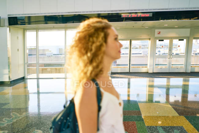 Seitenansicht einer verschwommenen, lockigen Frau mit Rucksack, die in der hellen Flughafenhalle mit glänzendem Marmorboden wandelt — Stockfoto