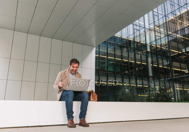 Cheerful successful man manager smiling and browsing laptop while sitting outside contemporary building on city street — Stock Photo