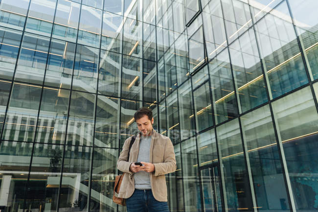 Angle bas de l'homme d'affaires heureux en utilisant smartphone tout en se tenant devant le centre d'affaires sur la rue de la ville — Photo de stock