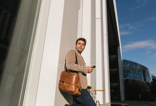 Angolo basso di serio imprenditore con smartphone appoggiato alla parete di un edificio moderno e guardando lontano nel centro città — Foto stock