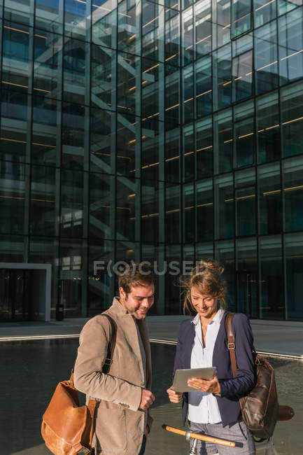 Fröhliche Männer und Frauen mit Fahrrad lächeln und schauen auf ein Tablet, während sie außerhalb des Bürogebäudes an der modernen Stadtstraße kommunizieren — Stockfoto