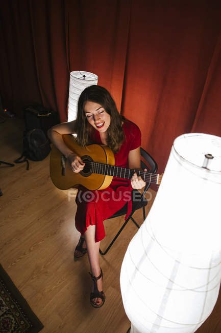 Von oben talentierte Frau in rotem Kleid singt und spielt Gitarre in warm beleuchteter Bühne neben weißer Lampe — Stockfoto