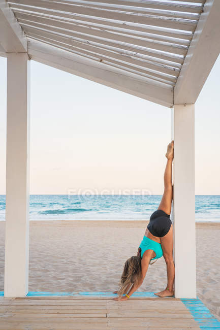 Side view of sportive power woman standing upside down on raising leg up on wooden stand on beach — Stock Photo
