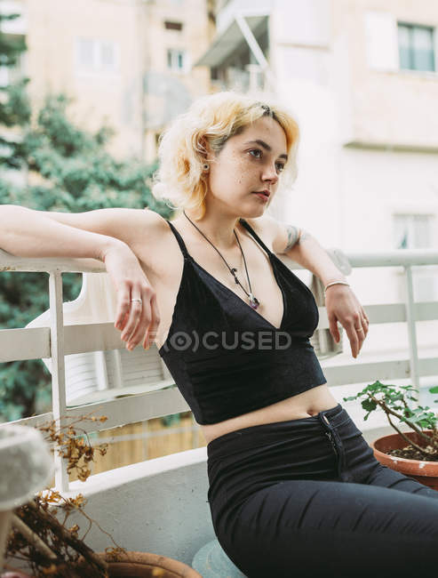 Mulher em roupas casuais pretas olhando para longe e contemplando enquanto sentado relaxado e inclinado sobre trilhos na varanda contra edifício residencial turvo — Fotografia de Stock