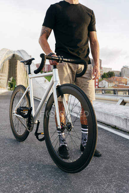 Immagine ritagliata di uomo che indossa camicia nera e pantaloncini beige in piedi con bicicletta bianca sul ponte con edifici della città su sfondo — Foto stock