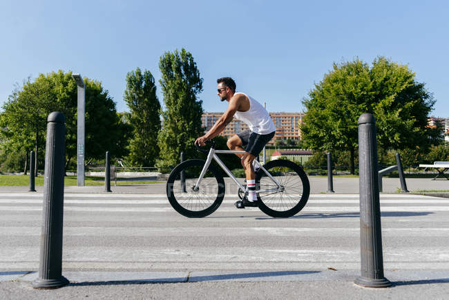 Vue latérale de l'homme athlétique portant des lunettes de soleil chemise blanche et short noir à vélo sur la route de la ville avec des arbres verts sur le bord de la route le jour d'été avec un ciel bleu — Photo de stock
