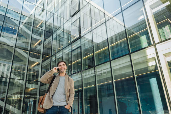 Низький кут щасливого бізнесмена тримає руку в кишені і використовує смартфон, стоячи за межами бізнес-центру на міській вулиці — стокове фото