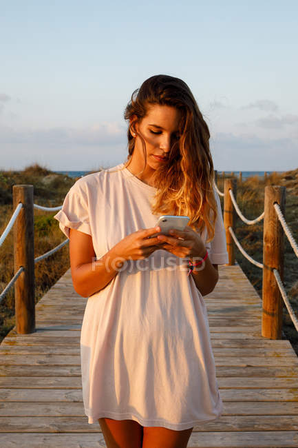 Pensiva donna dai capelli lunghi che naviga sul cellulare mentre cammina sul ponte sospeso alla luce del sole — Foto stock