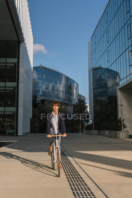 Delighted female entrepreneur smiling and riding bike on sunny day in downtown of modern city — Stock Photo