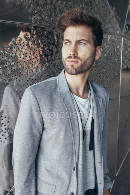 Hombre guapo de moda en chaqueta gris elegante y camiseta casual de pie y mirando hacia otro lado con la pared en el fondo - foto de stock