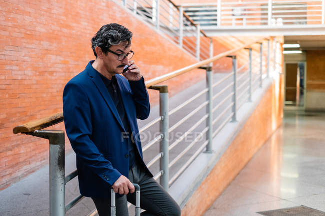 Empresario varón de edad en traje formal que tiene viaje de negocios con la maleta y llamando en el teléfono móvil con rampa de ladrillo en el fondo - foto de stock