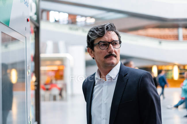 Bewildered handsome aged male entrepreneur in business style clothing and eyeglasses looking away on blurred background at shopping center — Stock Photo