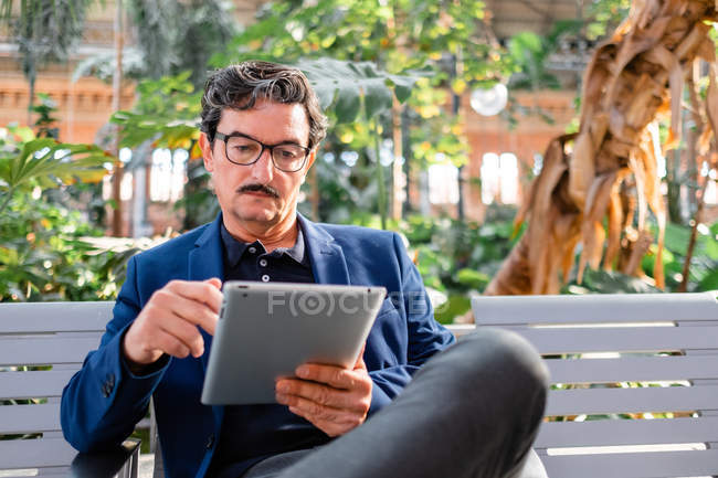 Smart elderly male remote worker in formal wear and trendy eyeglasses sitting with crossed legs on bench and working with digital tablet at park — Stock Photo