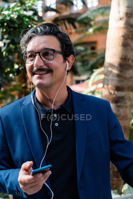 Cheerful elderly male owner in business style clothing having break and browsing mobile phone with headphones outdoors — Stock Photo