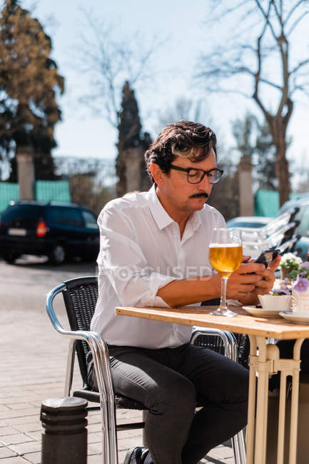 Пожилой бизнесмен в белой рубашке пишет смс на мобильный телефон, ожидая встречи на открытой террасе кафе со стаканом апельсинового напитка — стоковое фото